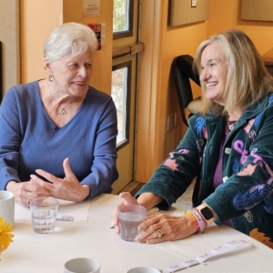 Two older women share a laugh over coffee at a lunch table, with a vase of fall flowers in the foreground.