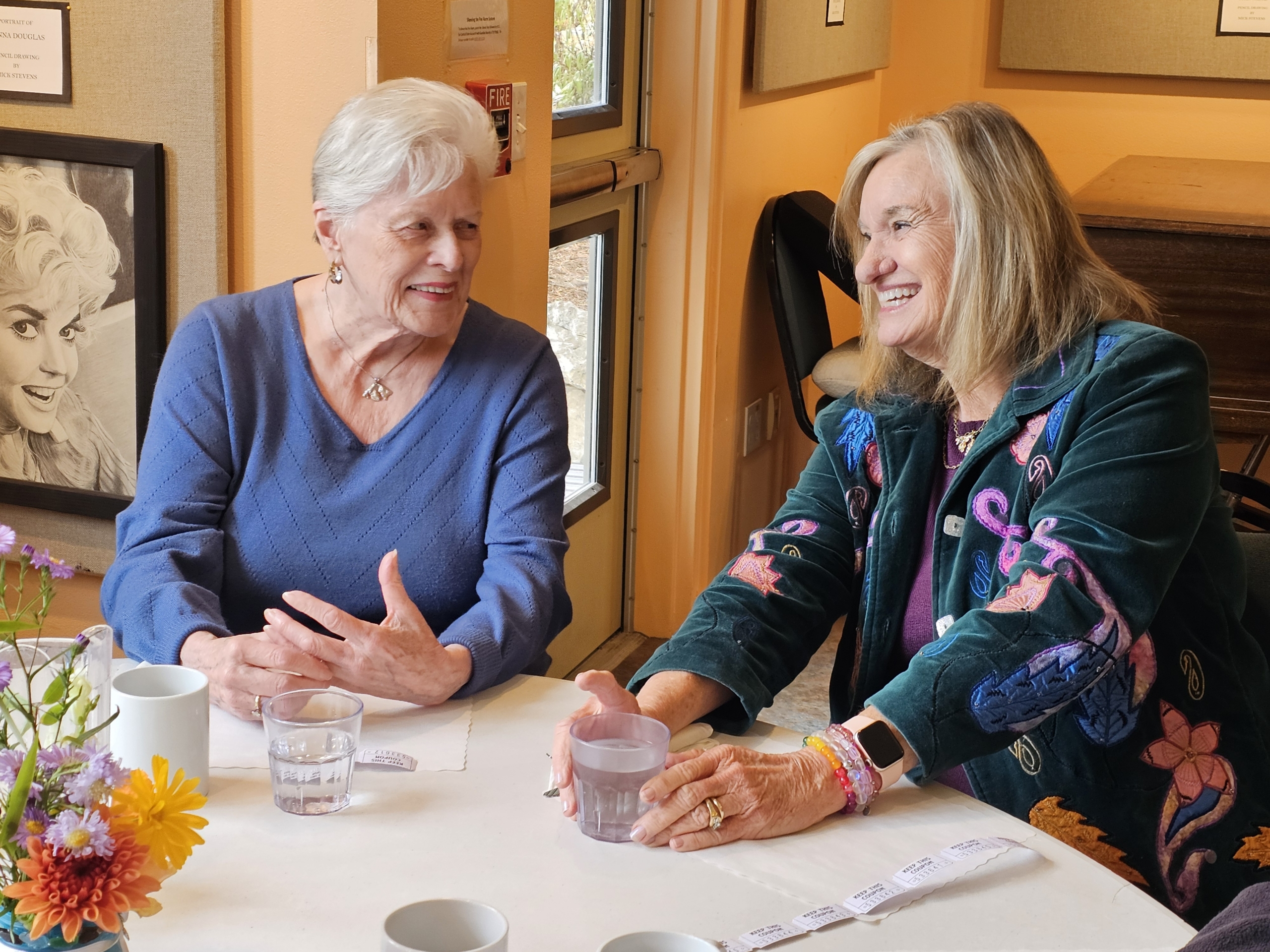 Two older women share a laugh over coffee at a lunch table, with a vase of fall flowers in the foreground.