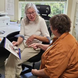 An older woman holding a Medicare booklet talks with a visitor, who points to a page, in a sunny office.
