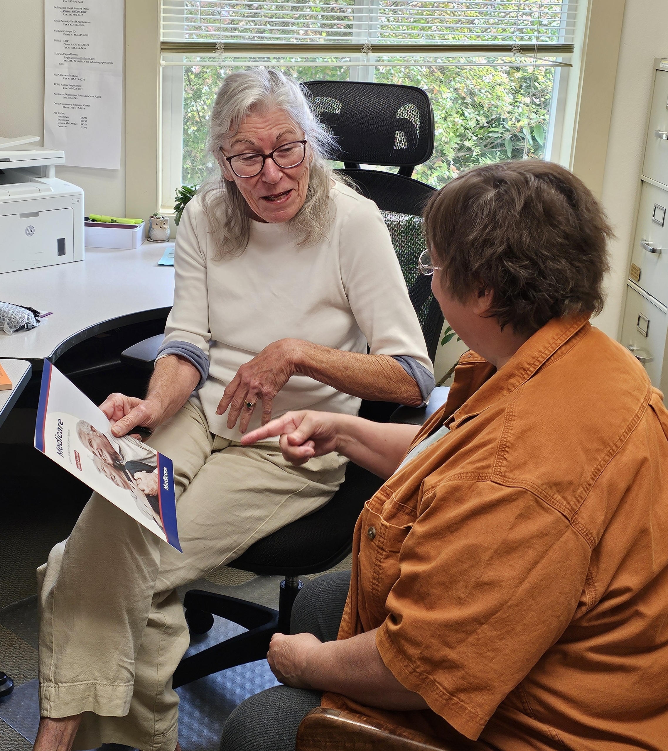 An older woman holding a Medicare booklet talks with a visitor, who points to a page, in a sunny office.