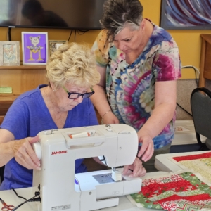 Two women work together at a sewing machine in a bright room at the Orcas Senior Center. One sits at a white Janome machine, guiding fabric under the needle, while the other leans in beside her, helping position the material. A festive red and green quilt top rests on the table nearby.