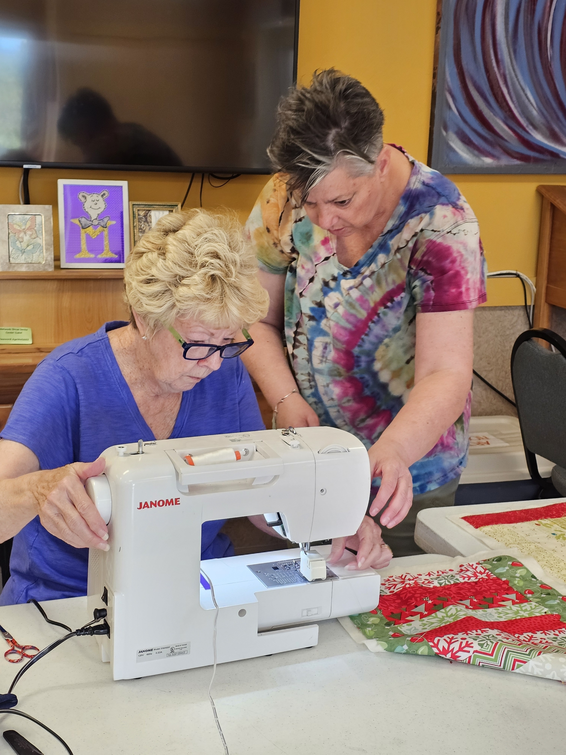 Two women work together at a sewing machine in a bright room at the Orcas Senior Center. One sits at a white Janome machine, guiding fabric under the needle, while the other leans in beside her, helping position the material. A festive red and green quilt top rests on the table nearby.