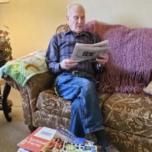 An older man in a striped shirt and jeans reads a newspaper on a floral sofa, with books and puzzle magazines in front of him.