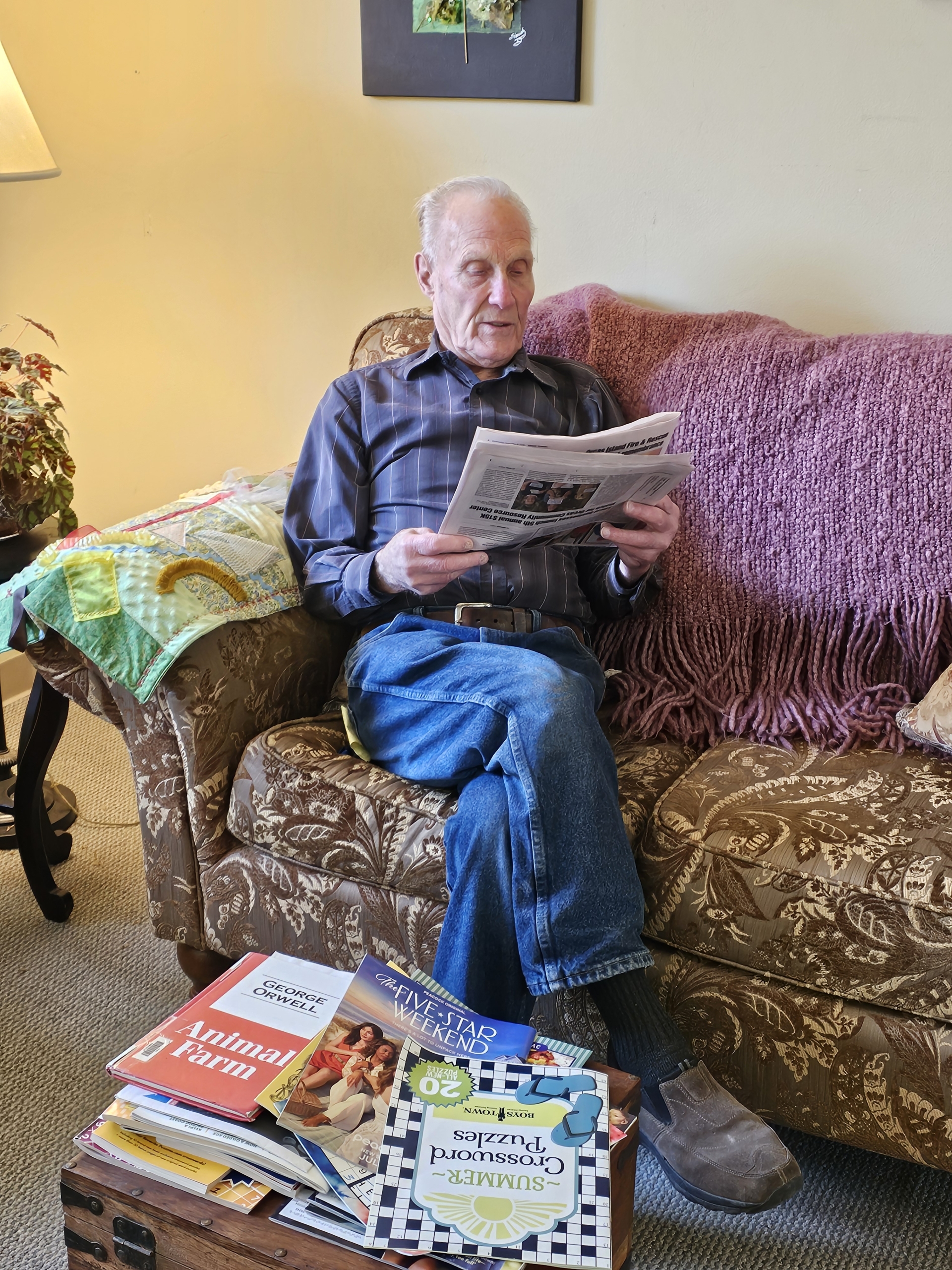 An older man in a striped shirt and jeans reads a newspaper on a floral sofa, with books and puzzle magazines in front of him.