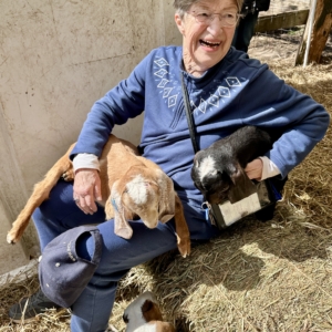 A smiling older woman sits in the straw of a sunny barn with two baby goats in her lap and a third goat kid at her feet.