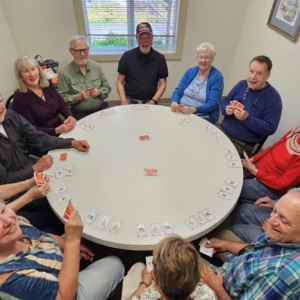 A group of smiling older adults sit around a large round table playing the card game Karma, holding red-backed cards with numbered cards laid face-up around the table's edge.