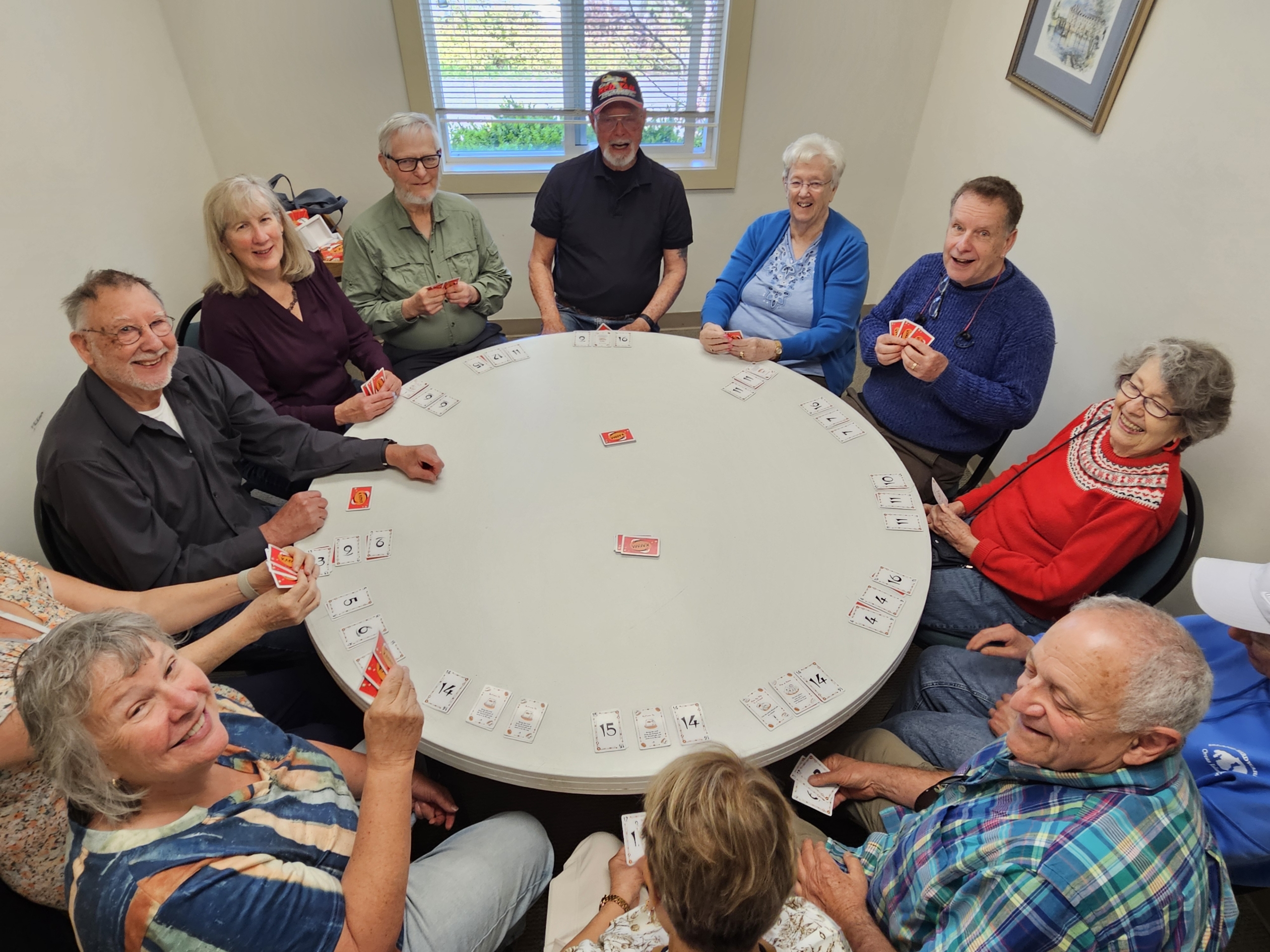 A group of smiling older adults sit around a large round table playing the card game Karma, holding red-backed cards with numbered cards laid face-up around the table's edge.