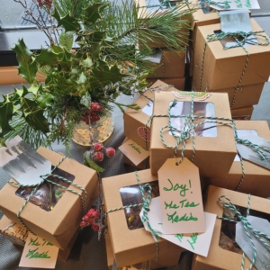 Kraft paper cookie boxes tied with green-and-white twine, with handwritten tags reading "Joy! The Tea Ladies," arranged beside a small vase of holly, pine, and red berries.